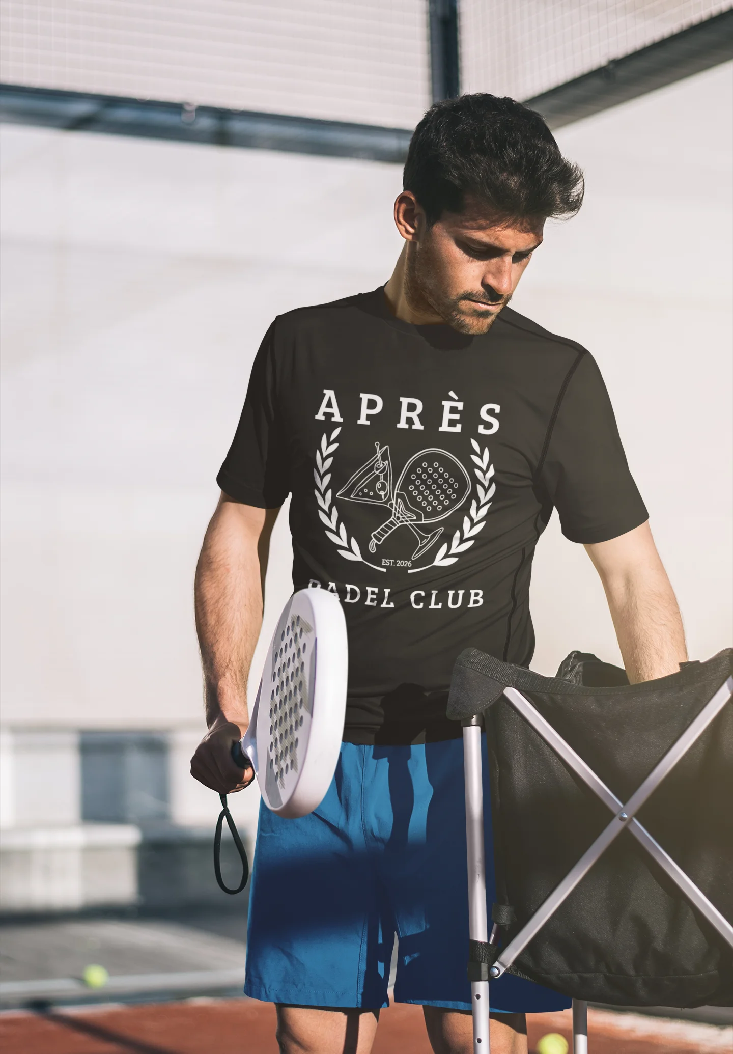 Padel Club Shirt Man holding a tennis racket and wearing an 'Apres Padel Club' t-shirt on a tennis court.