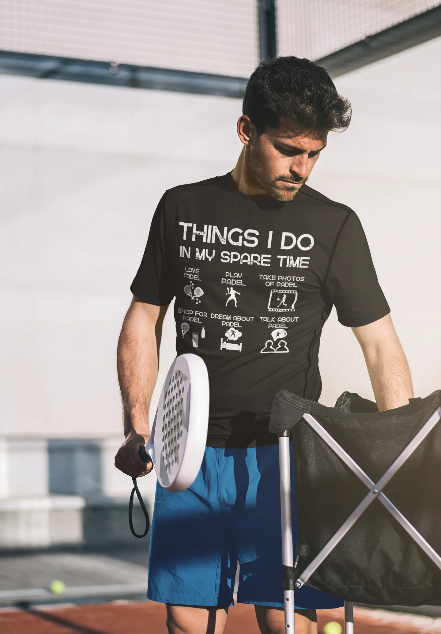 Man holding a paddle and a bag, wearing a black t-shirt with text, on a sports court.