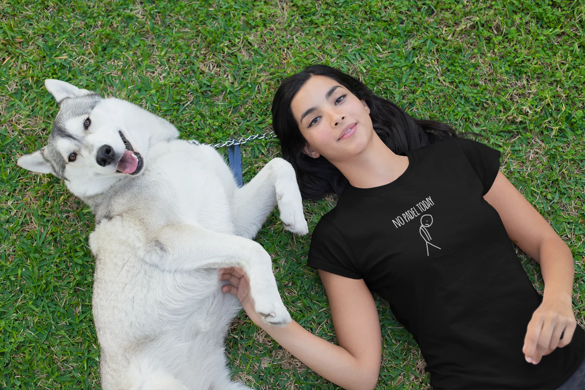Woman lying on grass with a white dog, wearing a black t-shirt with text and logo.
