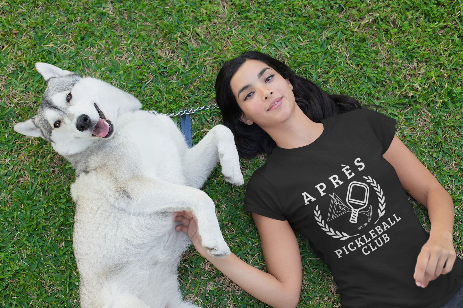 Funny Pickleball Shirt Woman lying on grass with a white dog, wearing a black t-shirt with 'Après Pickleball Club' text.