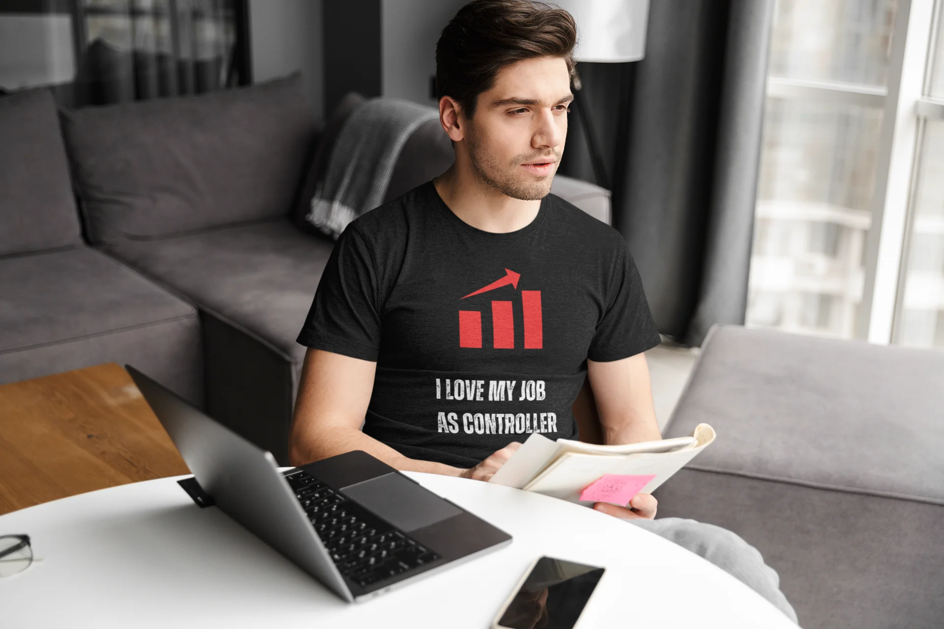 Finance Controller Shirt Man sitting on a couch with a laptop and tablet, wearing a black t-shirt with text.