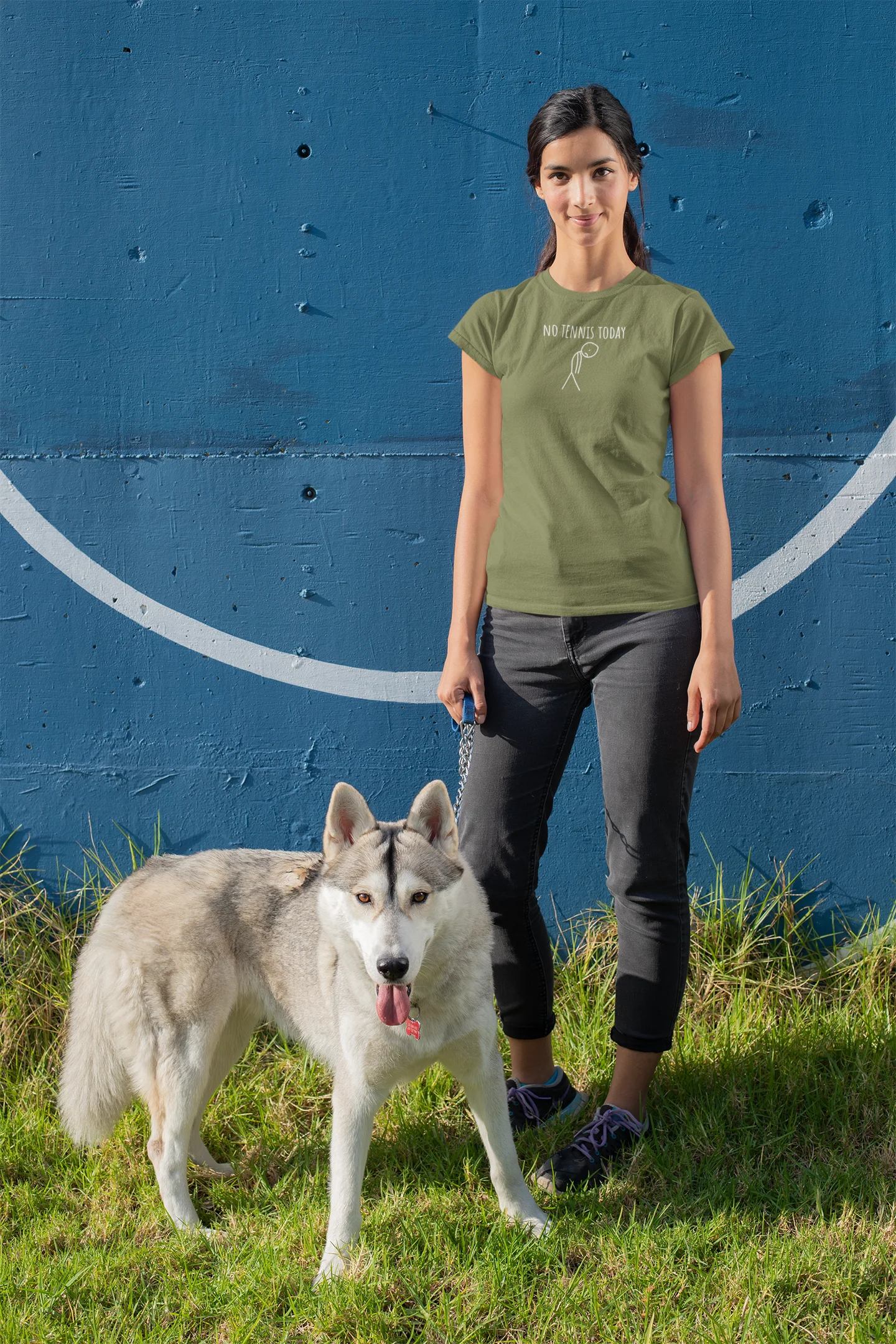 Woman in green t-shirt and dark pants standing next to a gray and white dog on grass with a blue wall background