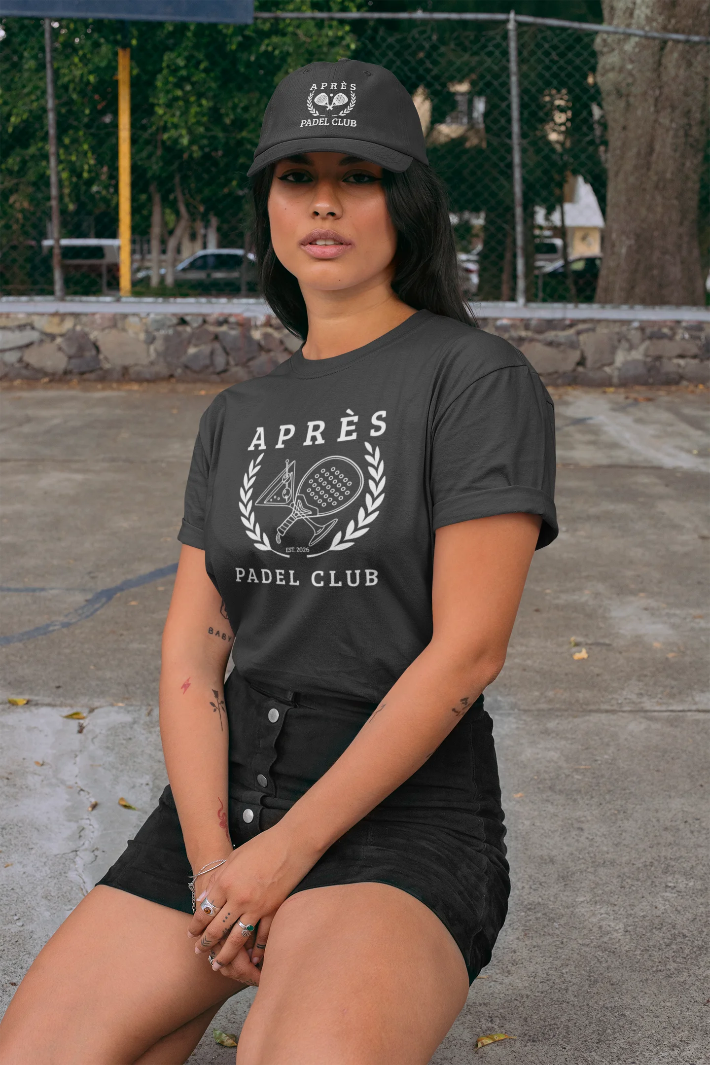 Padel Cap Woman wearing a black t-shirt with 'Apres Padel Club' text and logo, sitting outdoors.