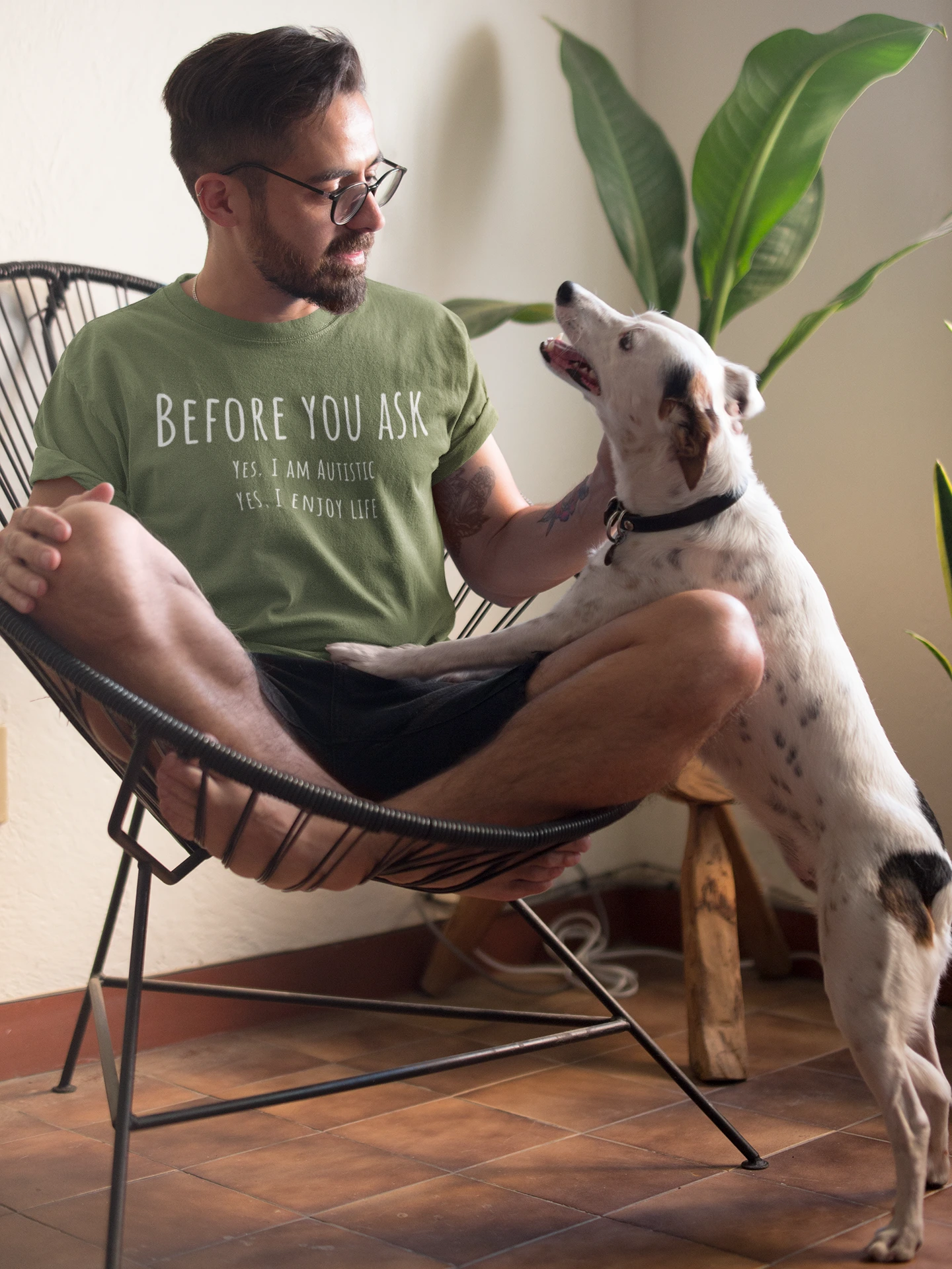 Man in a green t-shirt sitting on a chair with a dog, surrounded by plants.