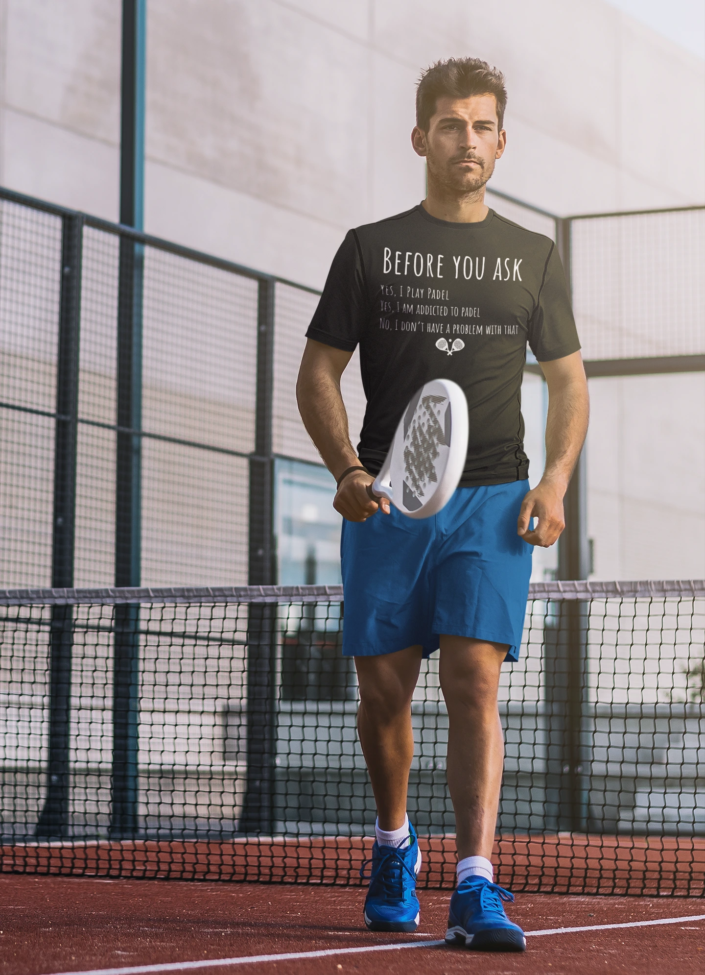 Man holding a padel on a tennis court wearing a t-shirt with text.