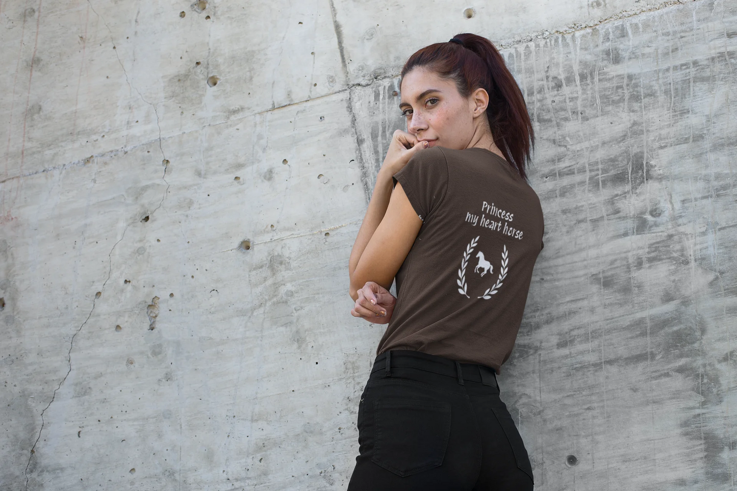 Woman wearing a brown t-shirt with a logo and text, leaning against a textured wall.