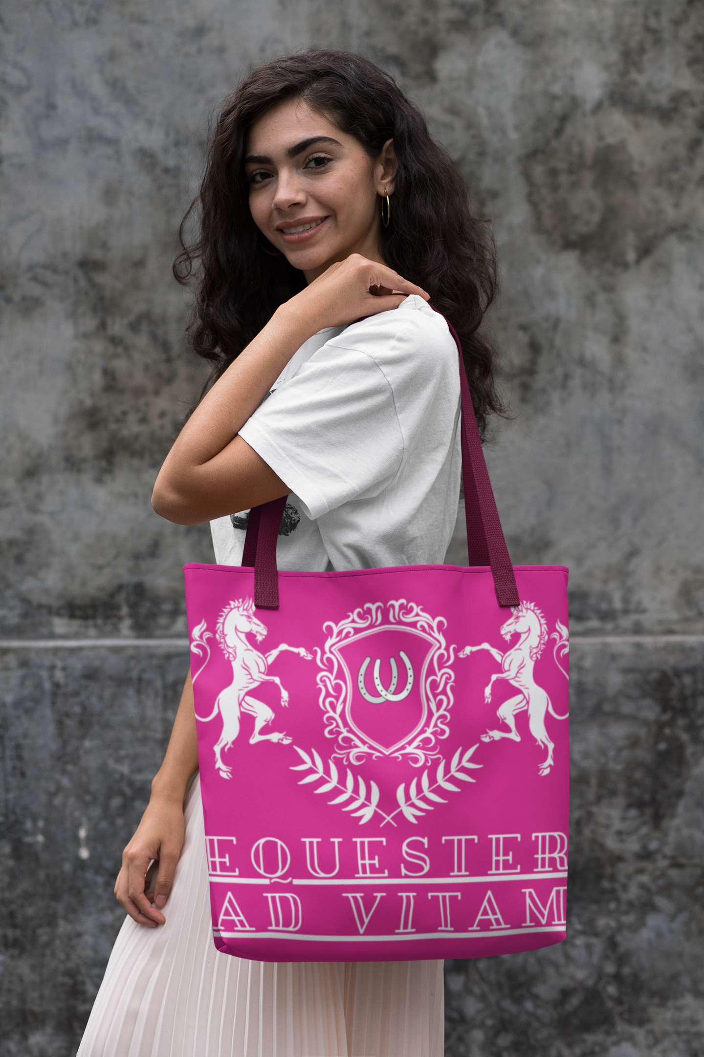 Woman holding a pink tote bag with a logo and text against a gray stone wall.