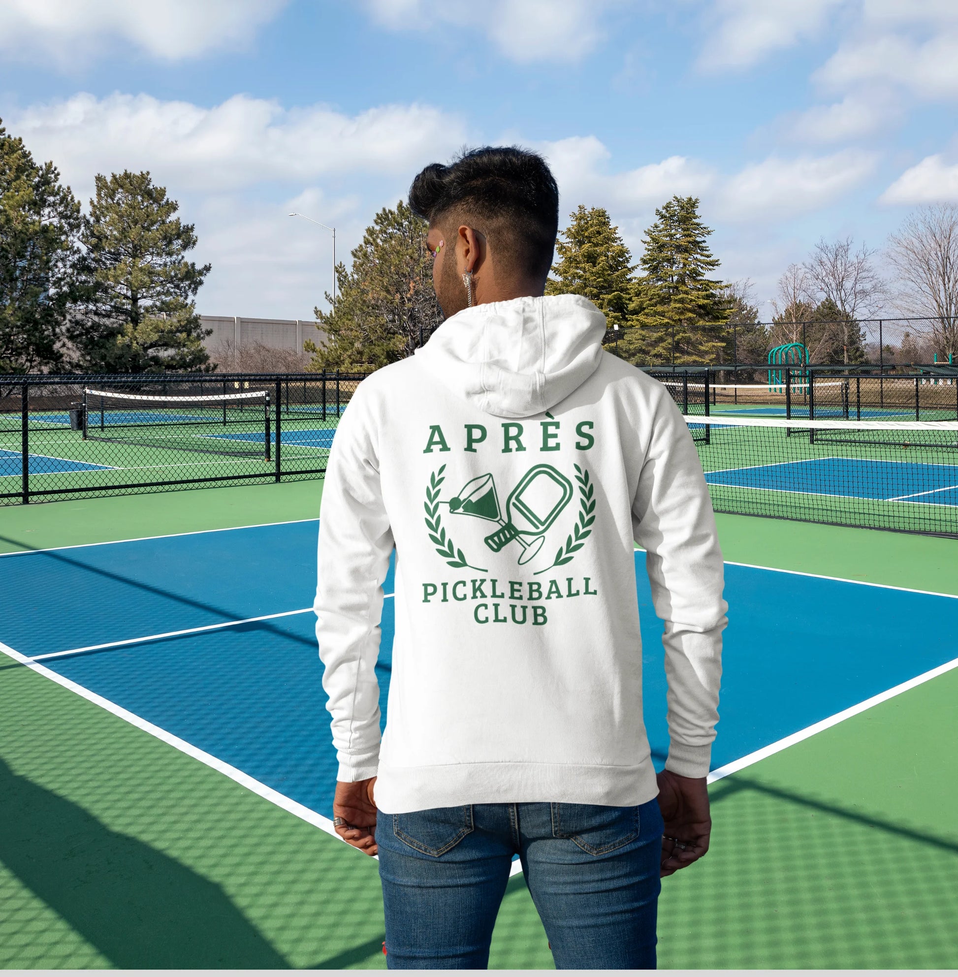 Warm up pickleball hoodie A man viewed from behind wearing a white hoodie with a green 'APRÈS PICKLEBALL CLUB' graphic, standing on a blue and green pickleball court.