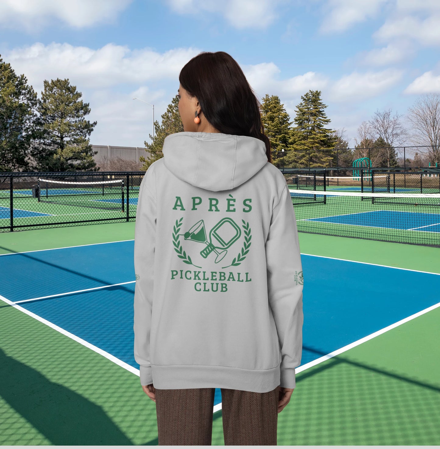 Warm up pickleball hoodie A person viewed from behind wearing a light gray hoodie with a green pickleball-themed graphic that reads 'APRÈS PICKLEBALL CLUB' on the back, standing on a blue and green pickleball court.