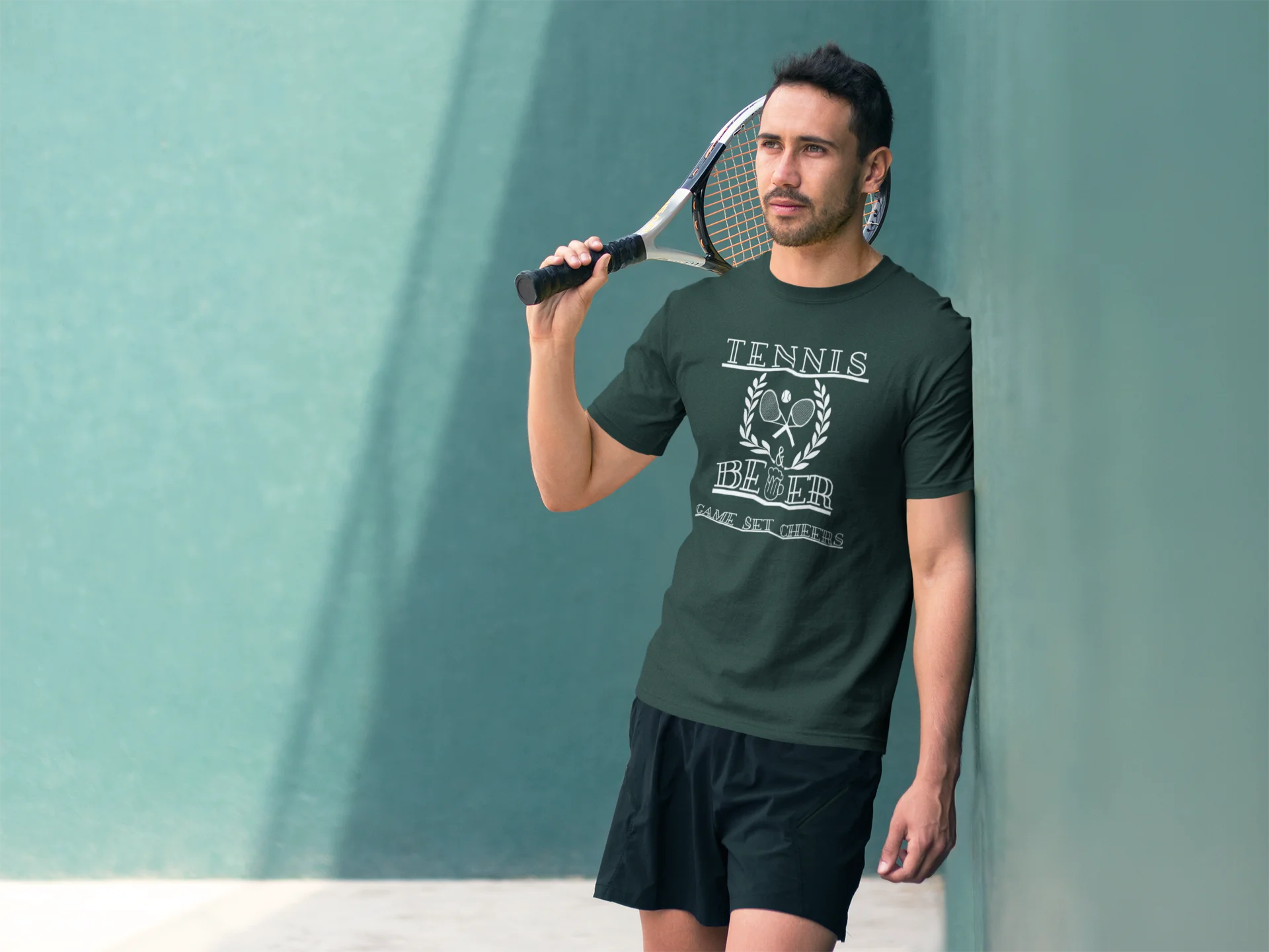 Man holding a tennis racket wearing a green 'Tennis Hero' t-shirt against a teal wall.