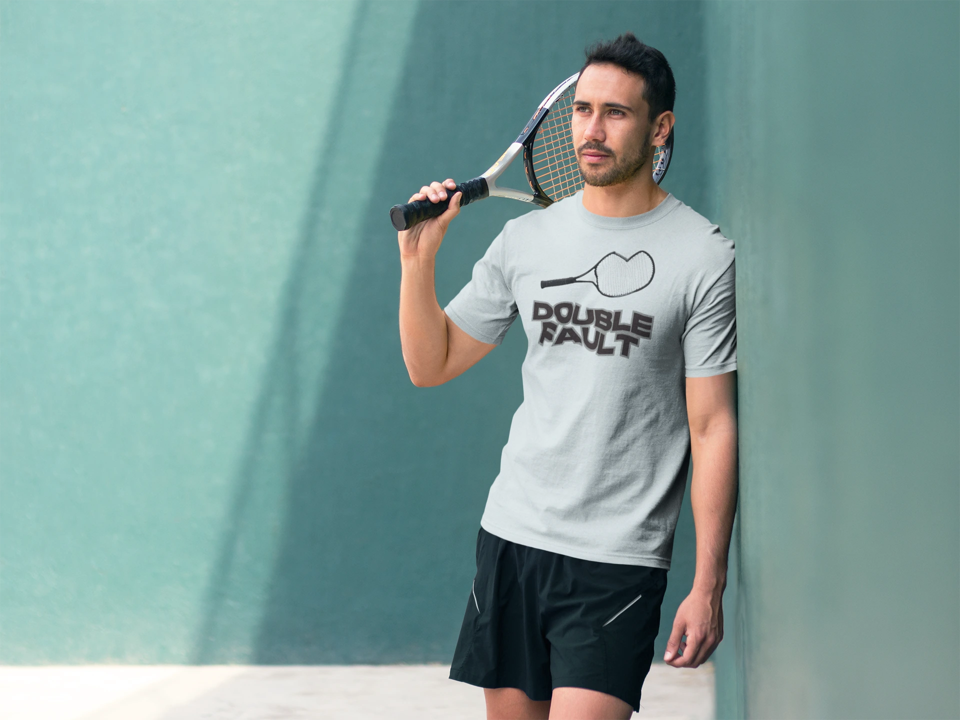 Double Fault Tee Man holding a tennis racket and wearing a t-shirt with 'Double Fault' text against a teal wall.